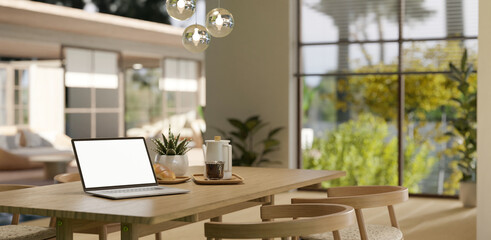 A white-screen laptop mockup on a wooden dining table in a spacious modern Scandinavian dining room.
