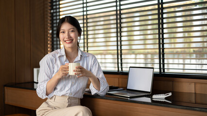 A beautiful Asian businesswoman is sitting at a table indoors with a coffee mug in her hands.