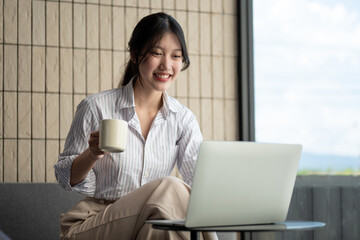 An attractive Asian woman is working on her laptop and sipping coffee, working remotely at a cafe.
