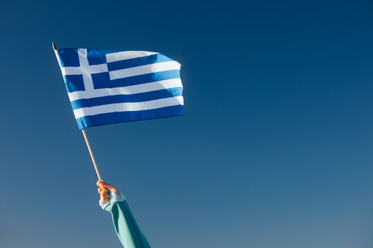 Hand Waiving a Greek Flag on a Blue Sky. Cheerful enthusiastic patriotic person displaying the symbol of Greece
 - Powered by Adobe