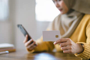 Young Muslim woman in a brown hijab shops online with credit card on mobile phone at cafe.