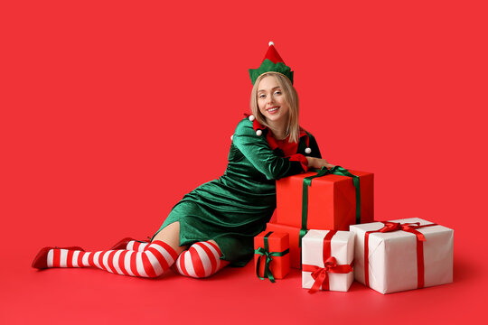 Happy young woman in elf costume with Christmas gift boxes on red background