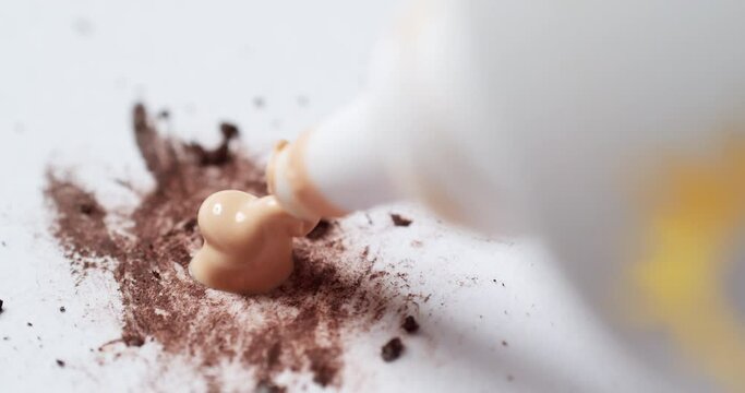 A close-up shot of a tube of liquid foundation concealer squeezed on a clean palette board to be mixed with some brown make-up tint.