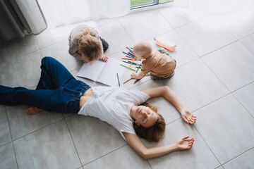 Tired mother lying on floor while her children drawing nearby. - Powered by Adobe