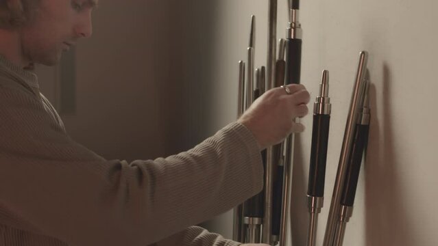 Side view medium shot of young Caucasian professional male gaffer choosing blowpipes for glassblowing in workshop