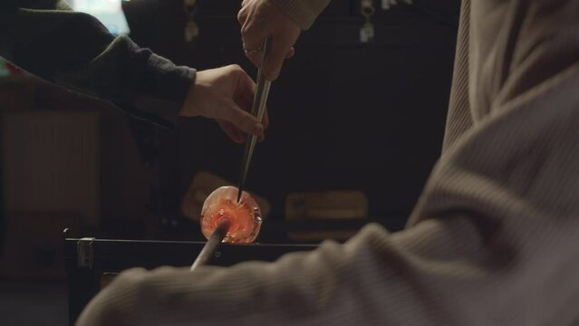 Cropped shot of unrecognizable male artisan glassblower shaping molten glass on end of pipe with tweezers and coworker helping him in workshop
