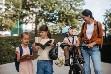 Mom picking up daughters from school with little son in bike seat. Schoolgirls telling mother about their day at school, walking in front of school building.