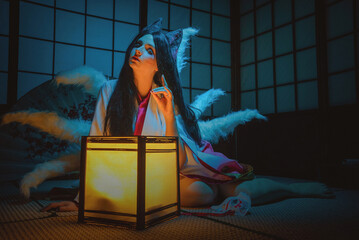 Fantasy young girl in the kimono and with fox ears in the light of floor lamps at the night sits on...