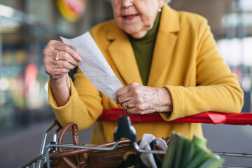 Elderly woman checking her receipt after purchase, looking at amount of money spent, ensuring all charges are correct. - obrazy, fototapety, plakaty