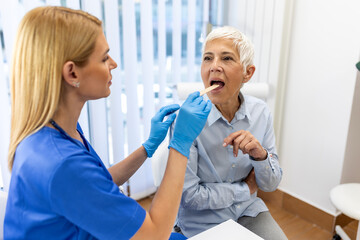 Obraz premium Senior patient opening her mouth for the doctor to look in her throat. Female doctor examining sore throat of patient in clinic. Otolaryngologist examines sore throat of patient.