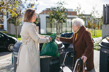 Woman helping elderly neighbor throw away trash into garbage can, waste container in front of their...