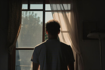 Rear view of adult man standing beside window with sunlight and shadow