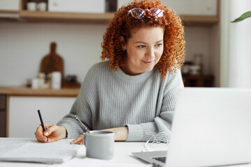 Portrait of happy female student watching webinar or tutorial studying online on laptop sitting at kitchen table, noting down after teacher or lecturer, looking at screen with interest - 677505709