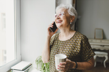 Side view portrait of happy smiling senior lady with gray hair in glasses holding cup of hot drink, enjoying phone conversation standing next to window in kitchen, talking to her best friend