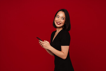 Naklejka premium Portrait of smiling woman using smartphone while standing isolated over red background