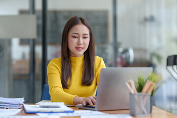 Happy young Asian woman smiling while working with laptop in office.