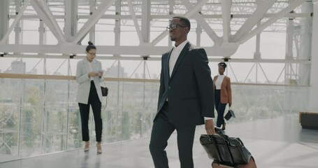 Dolly shot of elegant African American businessman walking in airport with suitcase while people with bags moving around. Modern city visible in panoramic windows.