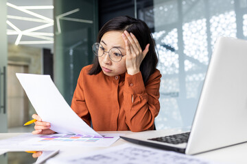Tired young chinese woman wearing glasses sitting at desk in office with pile of papers in hands,...