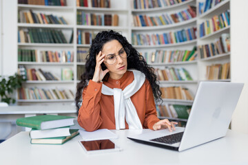 Tired young hispanic female teacher sitting at desk in front of laptop in school room holding hand on head, overwork in office headache, online learning concept.