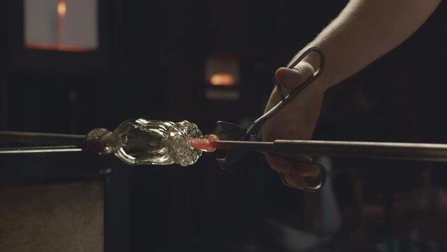Cropped shot of unrecognizable gaffer using stainless steel shears and spatula while molding shape of elegant transparent glass bottle produced with glassblowing technique in workshop