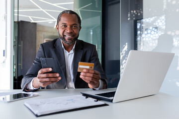 Senior African American man in suit sitting at desk in office. He is holding a credit card and a phone. Successfully paid online purchases, orders, bills. Happy looking at camera. - obrazy, fototapety, plakaty