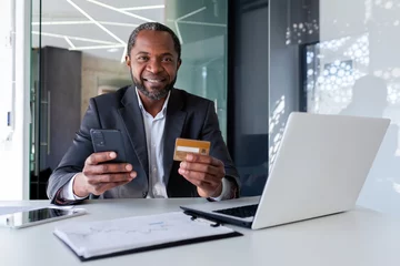 Foto op Aluminium Senior African American man in suit sitting at desk in office. He is holding a credit card and a phone. Successfully paid online purchases, orders, bills. Happy looking at camera. © Liubomir