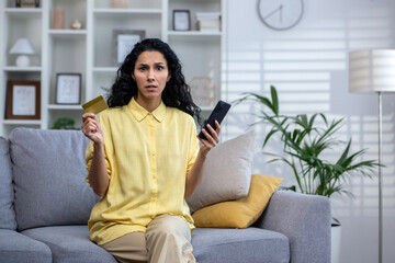 Upset hispanic curly haired girl sitting in light living room on sofa holding card and phone, hindu woman frustrated because of failed payment, fraud in online store, looking at the camera.
