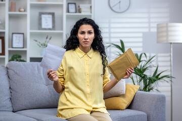 Annoyed curly haired hispanic woman sitting at home on sofa in living room holding open envelope. A cute brunette is upset about the news, losing the lottery, bad news.