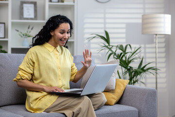 Happy hispanic woman sitting on sofa in bright living room with laptop on lap, video calling, distance learning, work meeting from home.