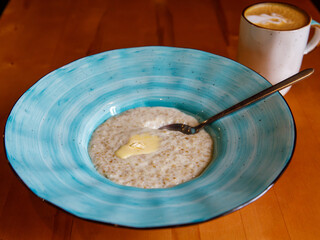 Healthy breakfast oatmeal porridge in blue bowl with spoon and cup of coffee on wooden table. Warm porridge oats with butter, vegan vegetarian weight loss dieting breakfast food. Close up