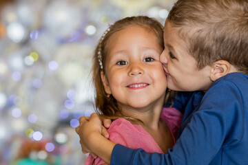 Obraz premium Cute boy standing by Christmas tree, hugging and kissing little sister. Merry Christmas and happy holidays