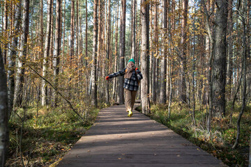 Fototapeta premium Overjoyed glad smiling middle-aged woman with shining of joy and happiness glance having rest in scandinavian pine forest reserve, hiking, enthusiastically jumping of pleasure on wooden pathway