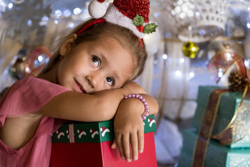 Adorable little girl sitting near Christmas tree, wearing Santa hat and holding present gift box. Merry Christmas and happy holidays