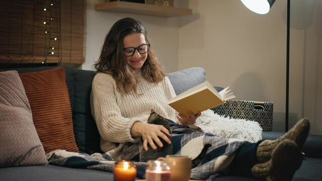 A young Caucasian woman reading a book sitting on the couch with her cute grey cat at home on a winter evening