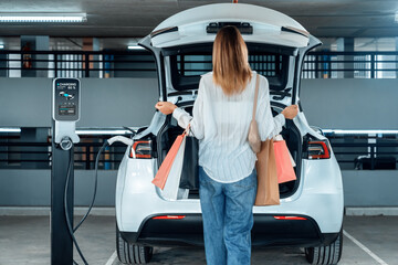 Young woman travel with EV electric car to shopping center parking lot charging in downtown city...