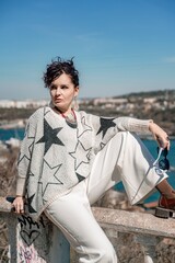 Woman walks around the city, lifestyle. A young beautiful woman in white trousers and a sweater sits on a white fence with balusters and overlooks the sea bay and the city.