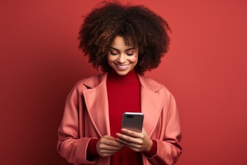 Portrait of beautiful positive girl with Afro haircut in pink jacket typing text message on smartphone, enjoying online communication, using app, chatting. Isolated on red studio background.