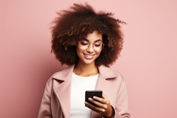 Fototapeta na wymiar Portrait of beautiful positive girl with Afro haircut in pink coat typing text message on smartphone, enjoying online communication, using app, chatting. Isolated on pink studio background.