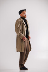 Stylish bearded man in flat cap, eyeglasses and trench coat standing on grey 