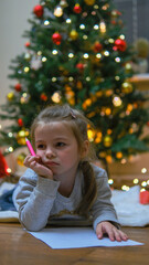 Fototapeta na wymiar Christmas routine. Portrait of a serious little girl in a red dress and Santa Claus hat lying on the floor under the Christmas tree writing a letter