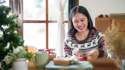 Young asian woman in sweater wrapping christmas gift and decorating with pine branch while preparing toys ornaments and presents to celebrate for christmas festive holiday and winter seasons at house