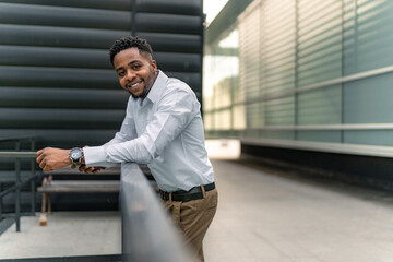 In a moment of respite, a charming Black businessman leans against a railing, absorbed in his phone...