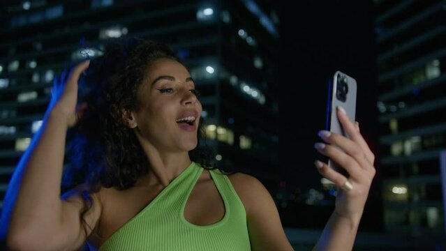 Happy flirting woman send video message to followers on social media. Young woman poses for selfie photos or video on smartphone camera. Night time screen lit image in city neon lights at downtown 4K