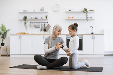 Fototapeta na wymiar Muslim female with daughter checking workout achievements on sports watch while sitting on yoga mat in kitchen. Mother and her girl taking advantages of home training using fitness tracker.