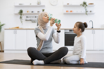 Beautiful adult arabian mom in sports clothes having sip of water while sitting with crossed legs on yoga mat in room. Little daughter girl posing looking at sporty mom having break training at home - obrazy, fototapety, plakaty
