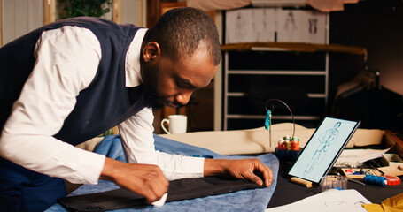 Male couturier marking fabric design to craft fashion items in tailoring atelier, using chalk to measure garment for needlework. Designer working on clothing line creation, workshop.
