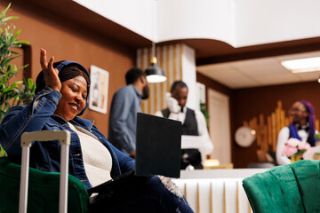 Happy smiling African American woman traveler sitting in hotel lobby having video call while...
