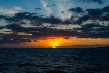 A dramatic orange sunset with colorful clouds and dark blue water from a boat on the Pacific Ocean off of the Island of Kauai in Hawaii, United States.
