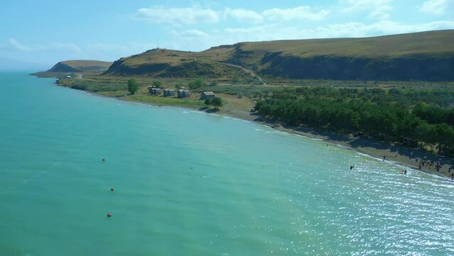 Aerial view of a large blue azure sea lake in clear sunny weather. People are swimming on the beach and pine trees are growing, boats are standing. Drone video, flying in an arc. Lake Sevan, Armenia.