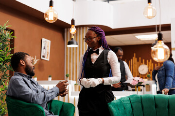 Smiling friendly African American woman hotel employee in uniform talking and interacting with happy guest in lobby. Generous rich man traveler giving cash tip to waitress girl at resort - obrazy, fototapety, plakaty